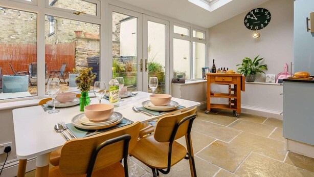 A dining area with a table set for four, a clock on the wall, and a view of the outside through large windows. - 2 Bed in Richmond (oc-90435) (Richmond)