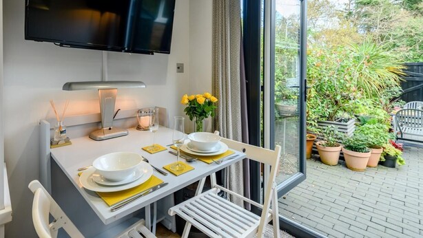 Ein Essbereich mit einem Tisch für zwei Personen, einem flachbildfernseher darüber und Blick auf den Garten durch eine Glastür.
