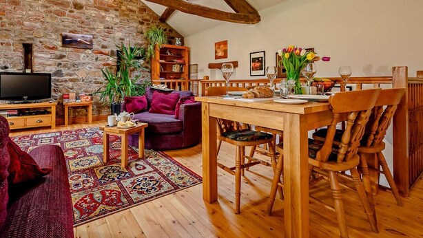 A cozy living room with a stone wall, wooden beams, a television, a sofa, a coffee table, and a dining area with chairs and a table set for two. - 2 Bed in Penrith (oc-90415) (Gamblesby)