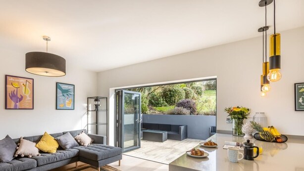 Una sala moderna con un sofá gris, un ventanal que da a un balcón y una zona de cocina con fruteros y una cafetera.