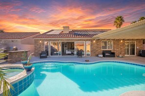 A residential swimming pool with a tiled deck, a patio area with outdoor furniture, and a brick house with a tiled roof.