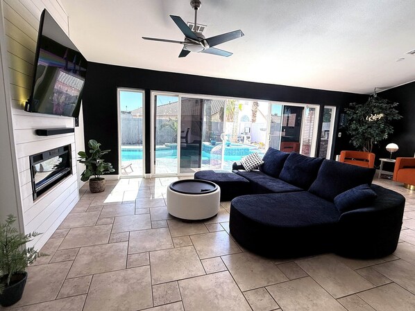 A modern living room with a sectional sofa, a flat-screen TV mounted on a white wall, a ceiling fan, and a view of a swimming pool through large glass doors.