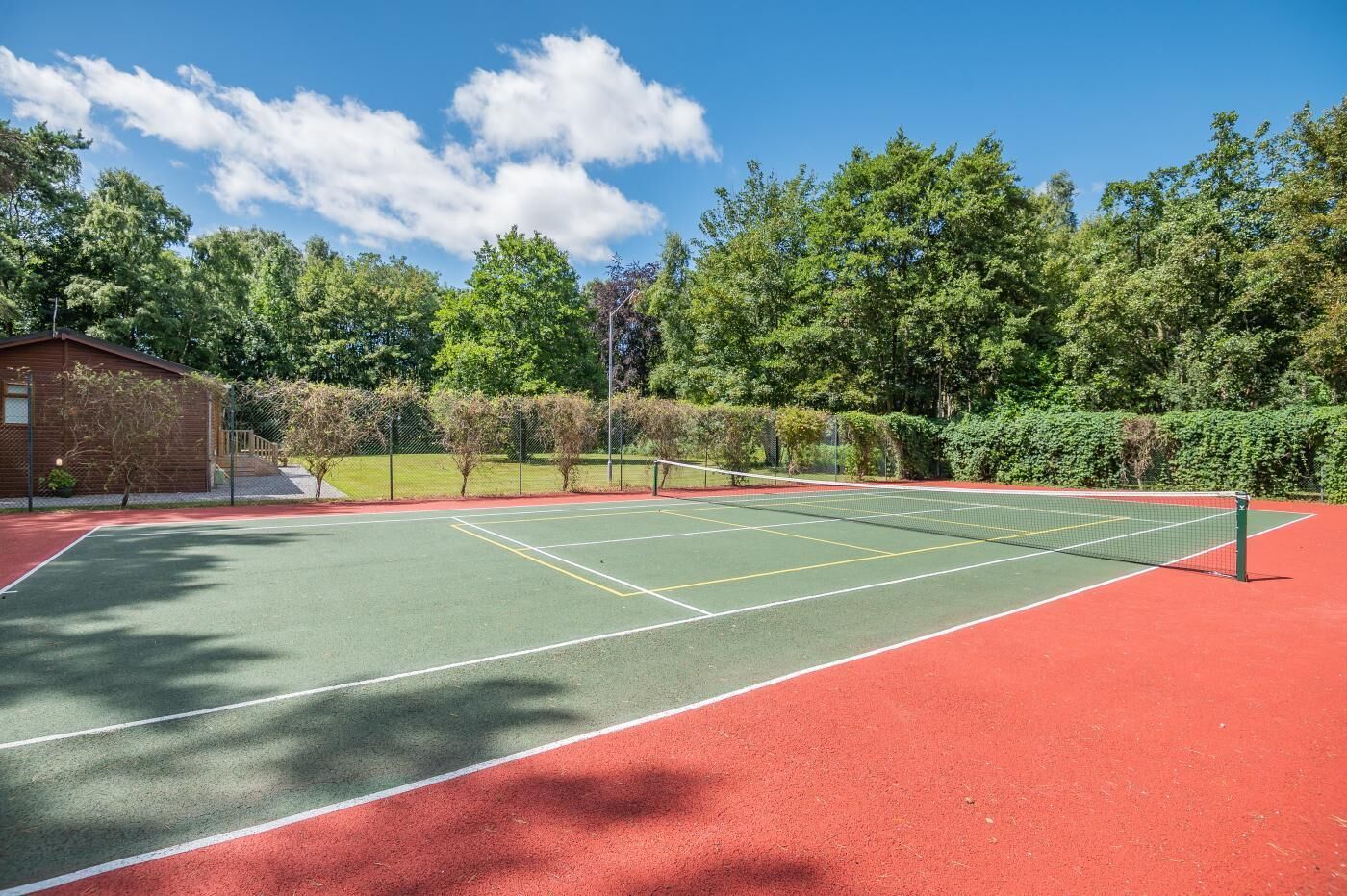 Una cancha de tenis con superficie roja y verde, rodeada de árboles y un pequeño edificio.