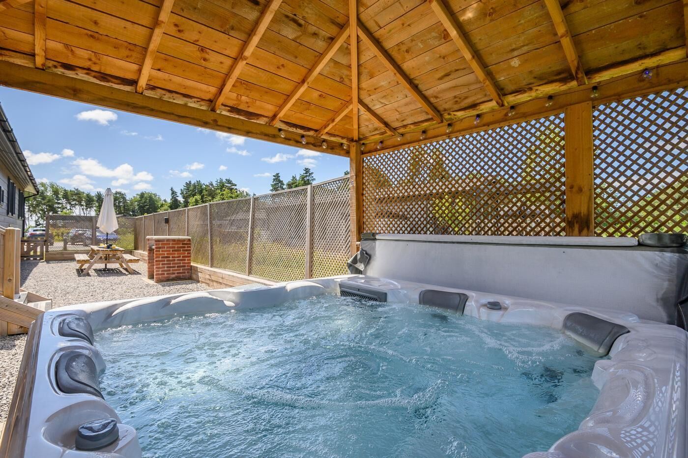 A hot tub under a wooden pergola with a fence and patio furniture in the background.