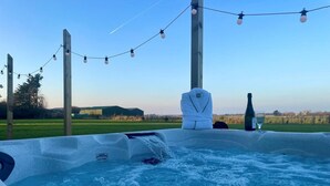 Ein Whirlpool mit Holzkonstruktion, einer Flasche und einem Glas, vor einer ländlichen Landschaft mit Lichterketten.