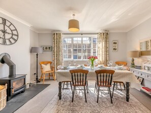 Ein Esszimmer mit einem für vier Personen gedecktem Tisch, einem Kamin und einem großen Fenster mit Vorhängen.