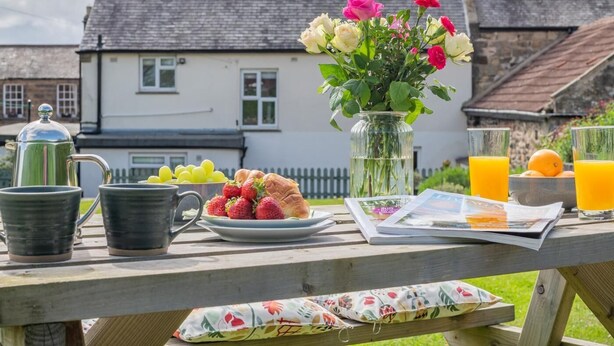 A picnic setup with a table, a vase of flowers, a newspaper, and a glass of orange juice. - 3 Bed in Belford (oc-90351) (Belford)