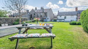 A picnic table with a floral cushion, a vase of flowers, and a spread of food and drinks in a grassy area with stone walls and a stone building in the background. - 3 Bed in Belford (oc-90351) (Belford)