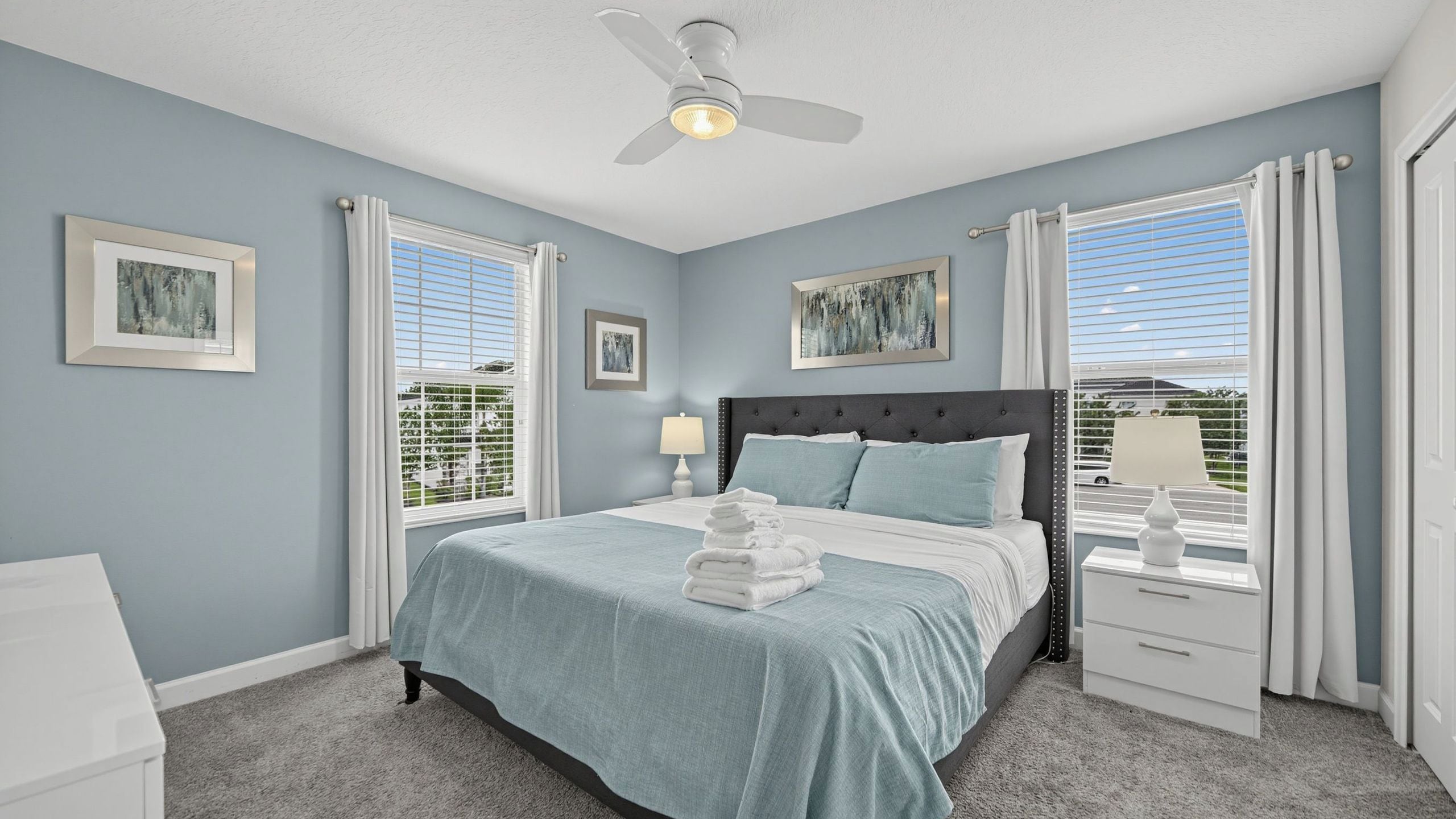 A bedroom with a bed, a nightstand, a lamp, a ceiling fan, and two windows with curtains.