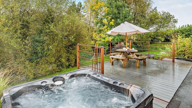 Un bain à remous entouré d’une terrasse en bois, une table de pique-nique avec un parasol et une clôture en métal.