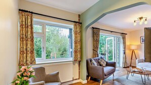 Un salon avec un canapé, un fauteuil et une table basse. Il y a deux fenêtres avec des rideaux, un radiateur et une lampe.