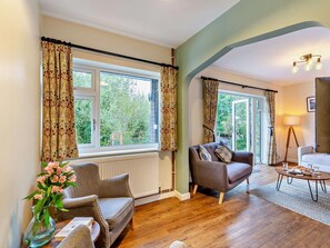 A living room with a sofa, armchair, and a coffee table. There are two windows with curtains, a radiator, and a lamp. - 3 Bed in Rhayader (oc-90282) (Saint Harmon)
