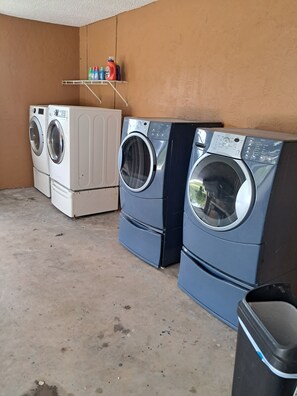 A laundry room with two front-loading washers and dryers, a shelf with cleaning supplies, and a trash bin. - Best Place in Avon Park (Avon Park)