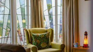 Um quarto com uma poltrona verde, uma almofada estampada, uma mesa de cabeceira de madeira com um candeeiro e vista através de uma janela com vitrais.