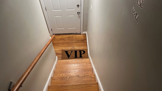 Eine hölzerne Treppe führt zu einer weißen Tür mit Fußmatte mit der Aufschrift 'VIP'.