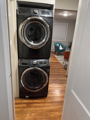 A stacked washer and dryer unit in a hallway with wooden flooring and a teal armchair in the background. - Cozy 2BR Apartment with Easy Access to DC Attractions (Washington)