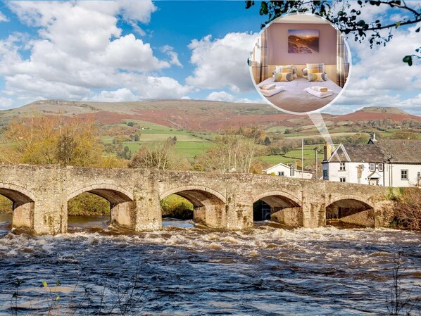 Un pont de pierre enjambe une rivière où l’eau coule, et on aperçoit, au loin, un bâtiment blanc ainsi que des collines verdoyantes.