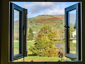 An open window with a view of a rural landscape, including a hill, trees, and a house.