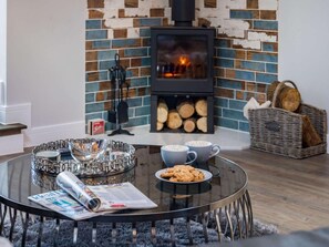 A cozy living room with a glass coffee table, a fireplace with logs, and a wicker basket. - 4 Bed in Axbridge (oc-90365) (Biddisham)