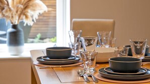 Et spisebord dækket med tallerkener, skåle, glas og bestik, med en vase med tørret pampasgræs på vindueskarmen.