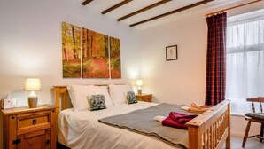 Ein Schlafzimmer mit einem hölzernen Bett, zwei Nachttischen mit Lampen, einem großen Gemälde mit Waldmotiv, einem Fenster mit Vorhängen und einem kleinen Stuhl.