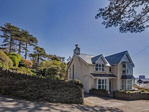 A two-story house with a stone wall, a pitched roof, and a garden with various plants and trees. - 5 Bed in Aberdovey (oc-90493) (Aberdovey)