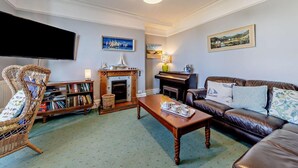 A living room with a fireplace, a sofa, a coffee table, a bookshelf, and a television. - 5 Bed in Aberdovey (oc-90493) (Aberdovey)