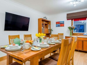 A dining area with a wooden table set for four, chairs, a television mounted on the wall, and a window with a view. - 3 Bed in Bathgate (oc-90491) (Fauldhouse)