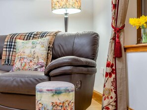 Ein braunes Ledersofa mit einer karierten Decke und einem floralen Kissen, ein kleiner runder Hocker mit floralem Muster, eine Stehlampe und ein Fenster mit einem blumigen Vorhang.
