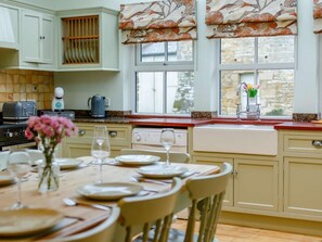 A kitchen with a dining area, featuring a stove, a coffee maker, and a window with curtains. - 4 Bed in Hexham (oc-90521) (Bardon Mill)