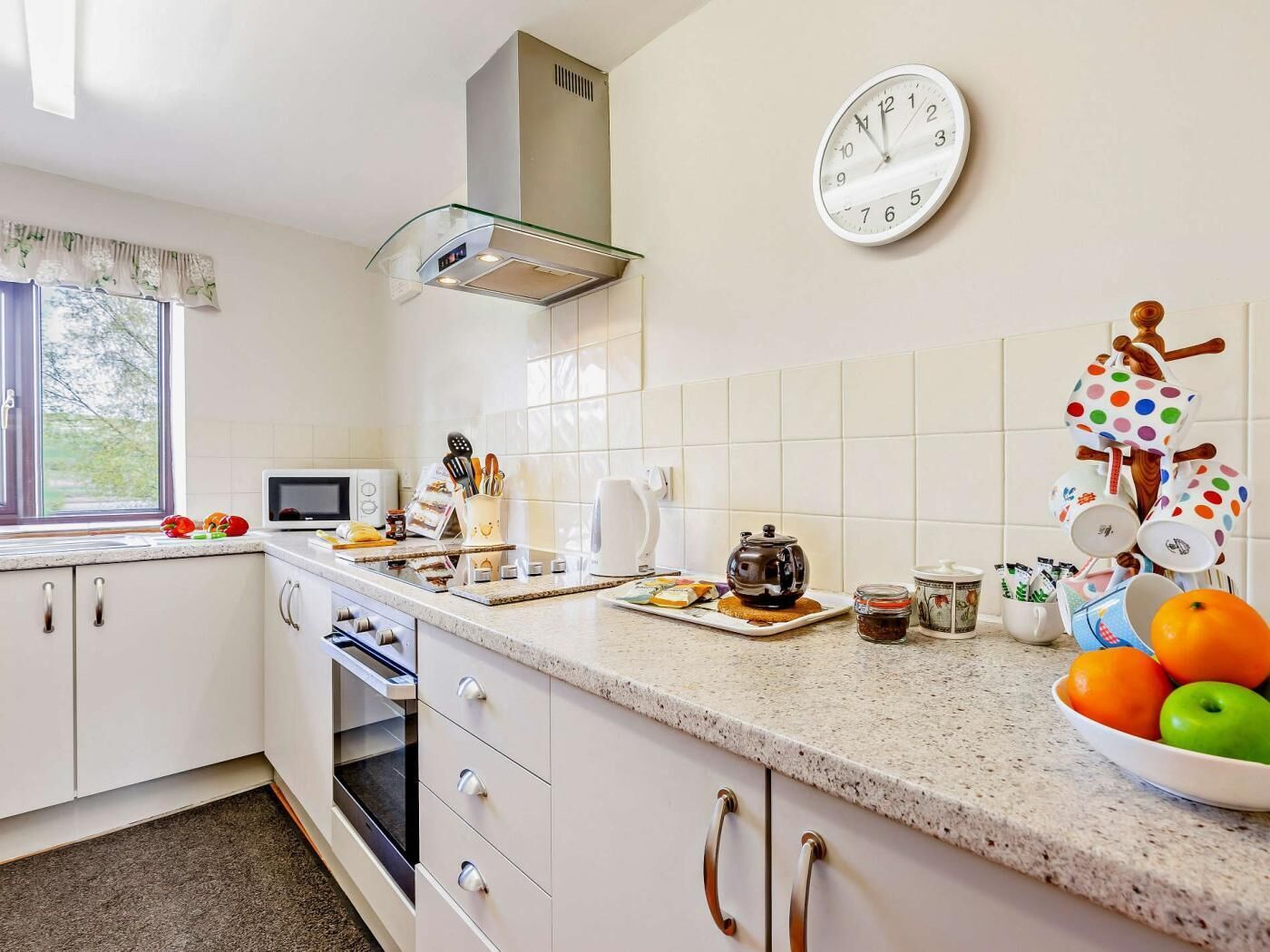 A kitchen with a microwave, tea pot, and a clock on the wall.