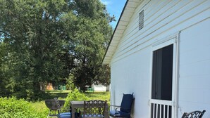 Um deck de madeira com mobiliário externo, um edifício branco com janela e uma grande árvore ao fundo.