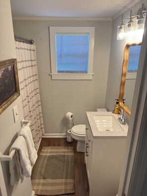 Un baño con inodoro, lavamanos, ventana con persianas, perchero para toallas y espejo.