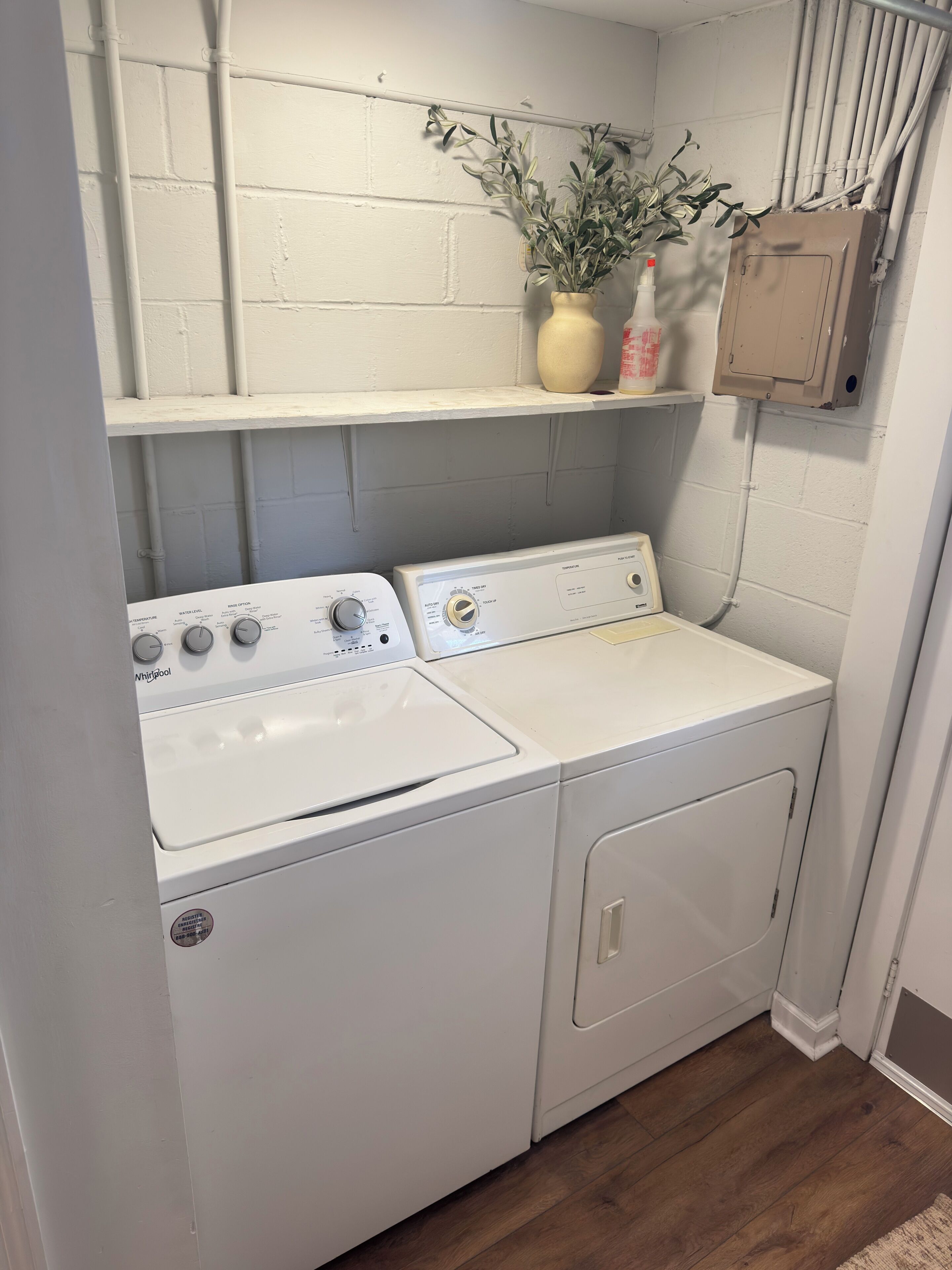 Ett tvättrum med två vita tvättmaskiner och torktumlare från Whirlpool, en hylla med en krukväxt och en flaska samt en beige elcentral.