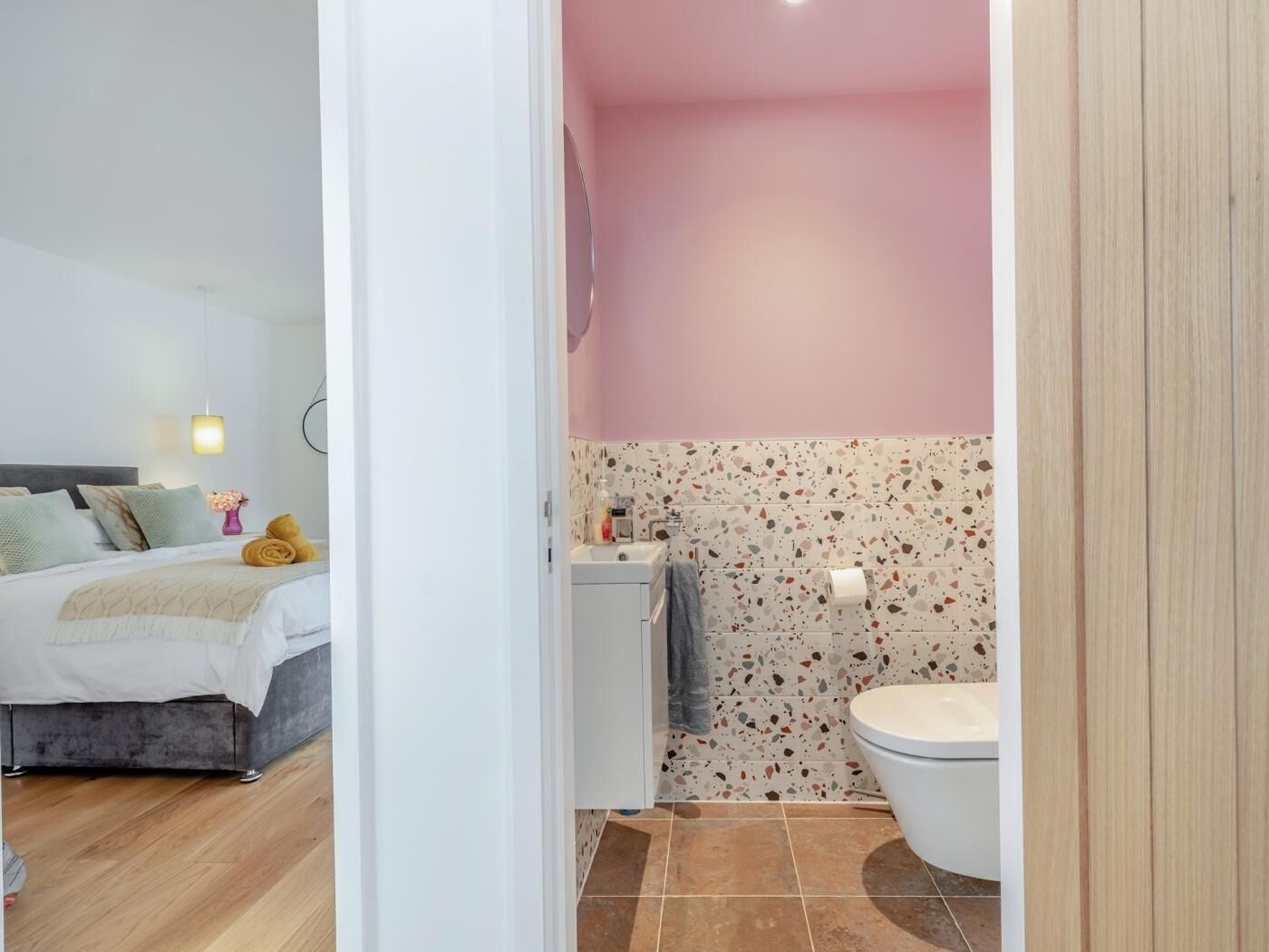 Et soveværelse med seng, natbord og lampe, samt et badeværelse med toilet, vask og bruser.