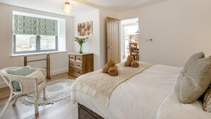 Ein Schlafzimmer mit einem Bett, einem Sessel, einer Kommode, einem Fenster mit gemusterten Vorhängen und einer Tür zu einem anderen Raum.