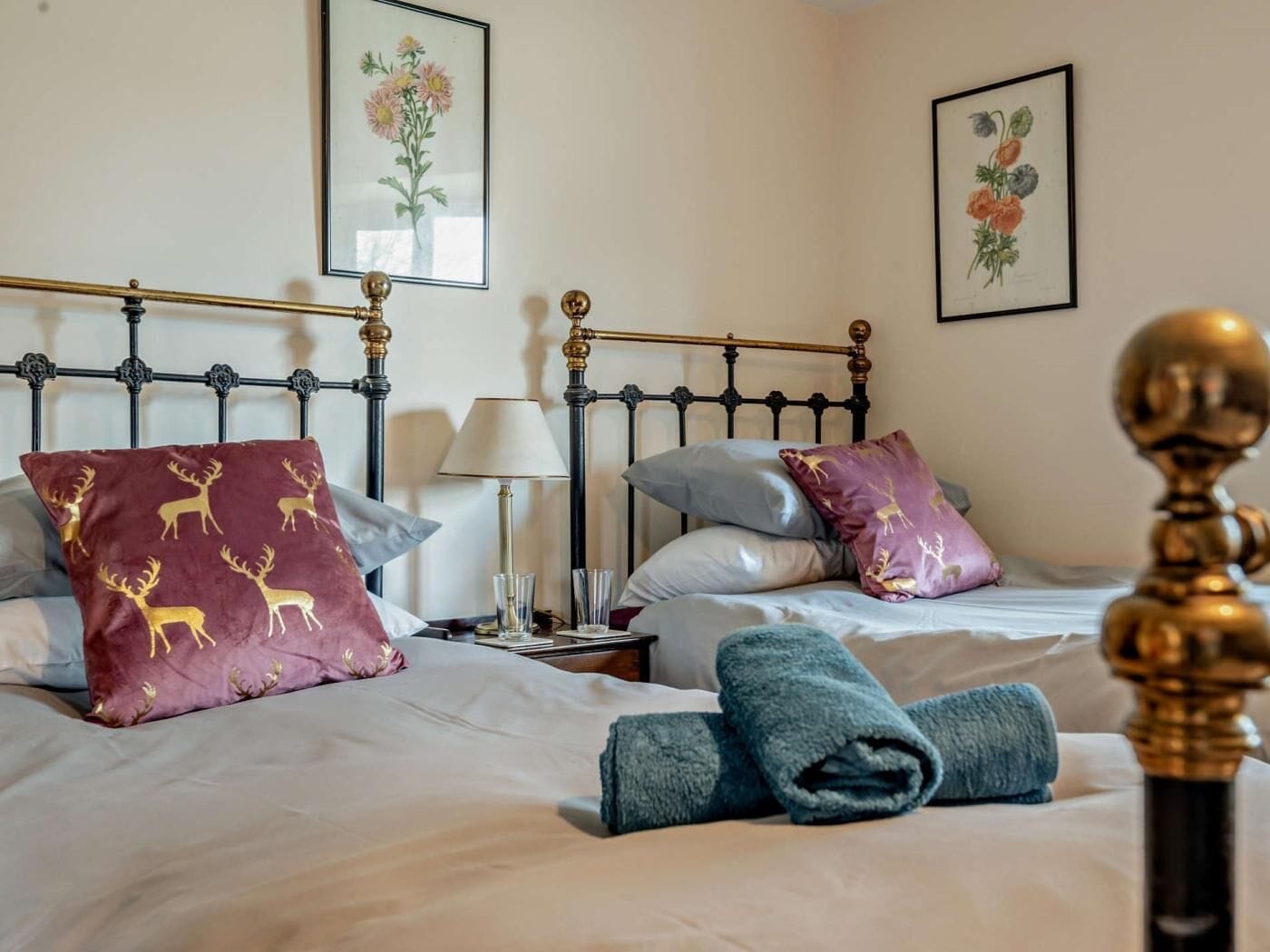 A double bed with a metal frame, two pillows with deer patterns, a blue towel, and a bedside table with a lamp.