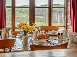 A breakfast table set with croissants, coffee cups, and a jar of jam, with a vase of daffodils in the center, overlooking a scenic view.
