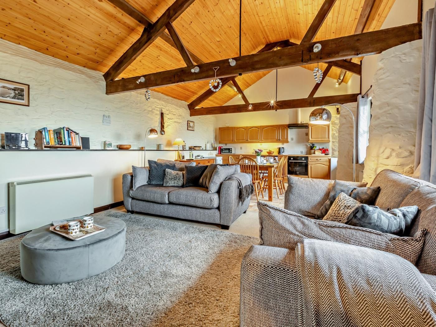 A spacious living area with a high wooden ceiling, a grey sofa, a wicker armchair, and a kitchen in the background.