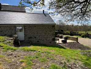 Un bâtiment en pierre avec un toit en pente, une porte et un petit jardin agrémenté d’un banc et de jardinières.