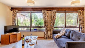 Un salon avec un canapé, un téléviseur, une table en bois sur laquelle se trouvent une bouteille et des verres, et une fenêtre avec des rideaux.