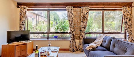 A living room with a sofa, a TV, a wooden table with a bottle and glasses, and a window with curtains.