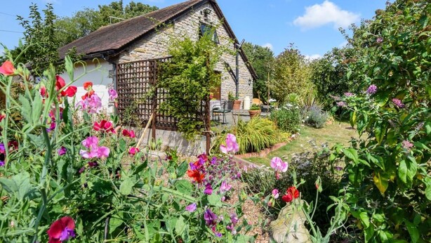 Une maison en pierre avec un toit en pente, entourée d’un jardin où poussent différentes espèces de plantes à fleurs.