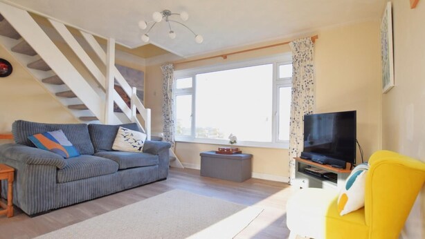 Ein Wohnzimmer mit einer Couch, einem gelben Sessel, einem Fernseher, einer Treppe und einem Fenster mit Vorhängen.