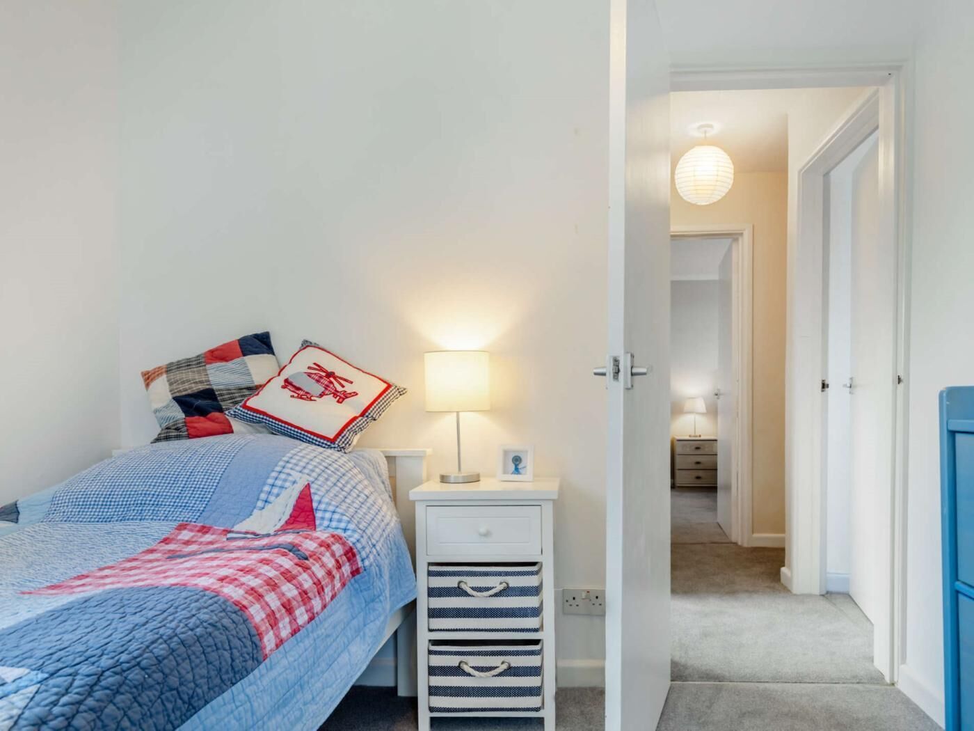 A bedroom with a bed, bedside table, lamp, and a view into a hallway.