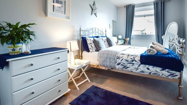 A bedroom with a bed, dresser, bedside table, lamp, potted plant, and a window with curtains. - 1 Bed in Whitby (oc-90699) (Fylingdales)