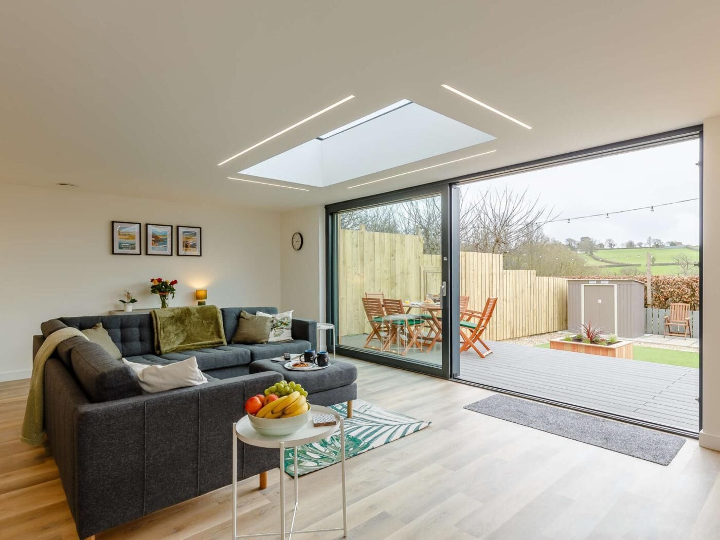 Un moderno salón con un sofá gris, una mesita con fruta y un ventanal que da a un patio exterior con terraza de madera y sillas.