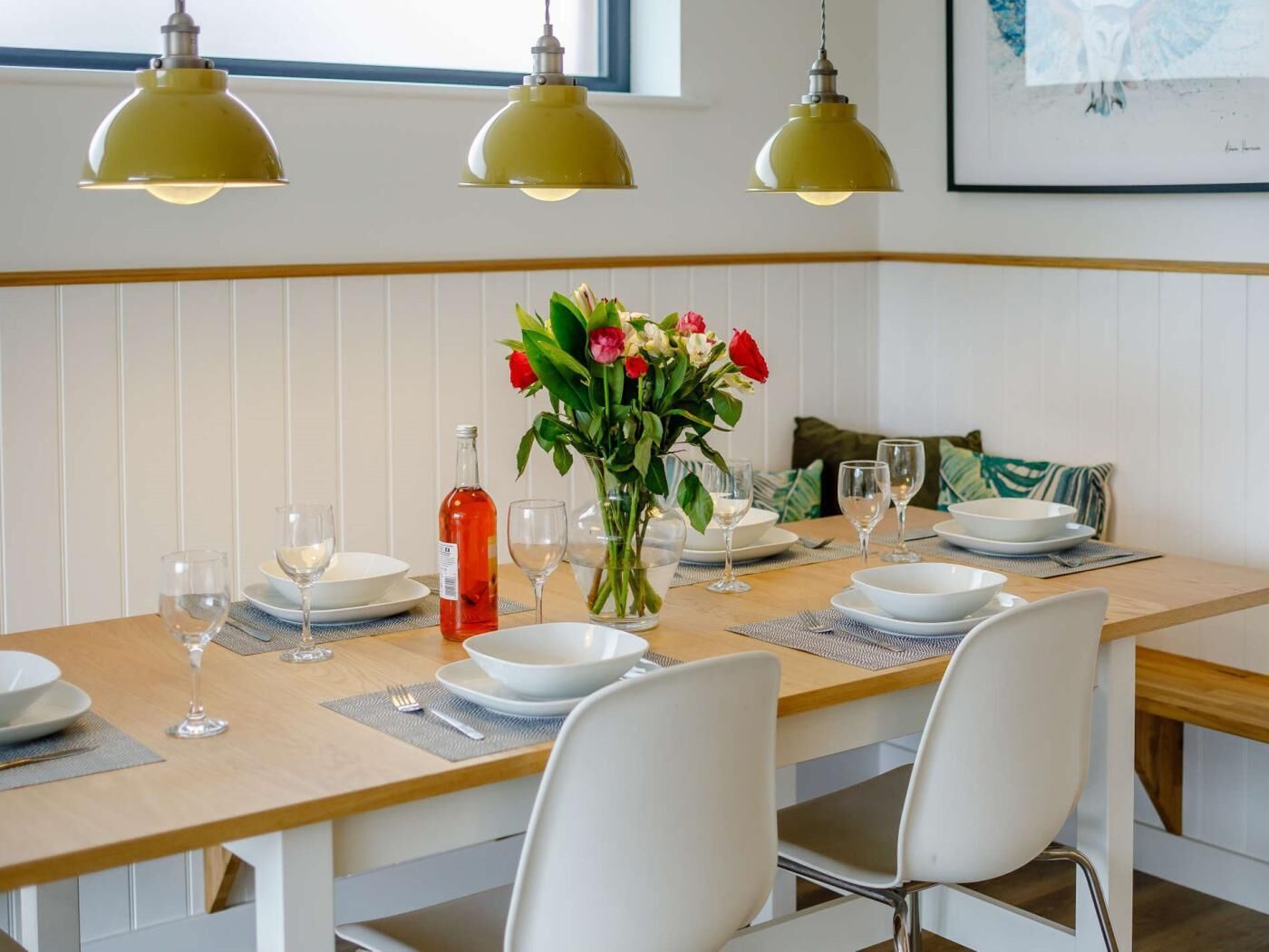 Une table à manger dressée avec de l’assiette blanche, des verres et une bouteille, et un vase de fleurs placé au centre.