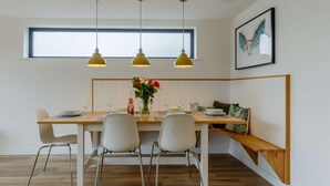 A dining area with a wooden table, four chairs, a vase of flowers, and three pendant lights. - 3 Bed in Holsworthy (oc-90721) (Holsworthy)