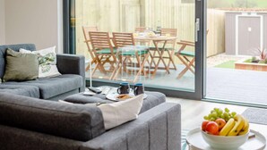 A living room with a grey sofa, a coffee table with a bowl of fruit, and a view of an outdoor patio with a wooden table and chairs. - 3 Bed in Holsworthy (oc-90721) (Holsworthy)